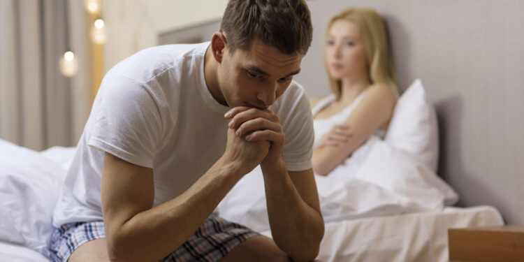 relationships, people, divorce and sexual problems concept - upset man sitting on bed with woman in bedroom